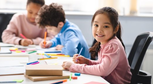 Three children drawing with crayons at a table, one girl looks out smiling